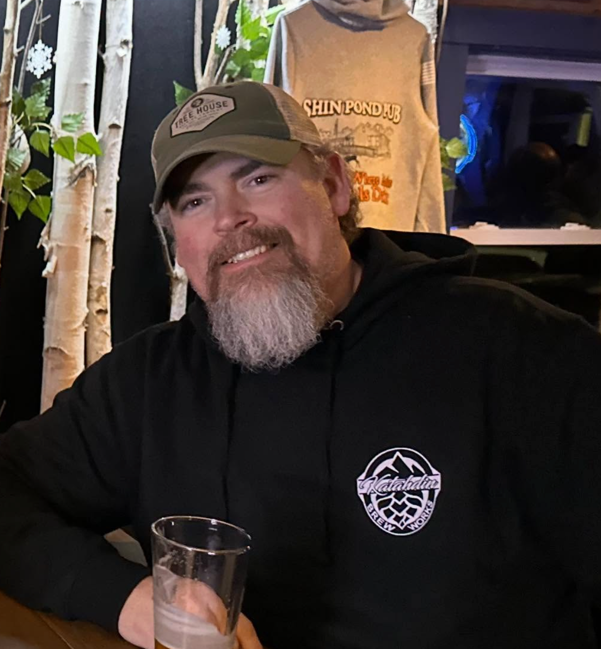 man sits smiling holding a beer with a wooded background